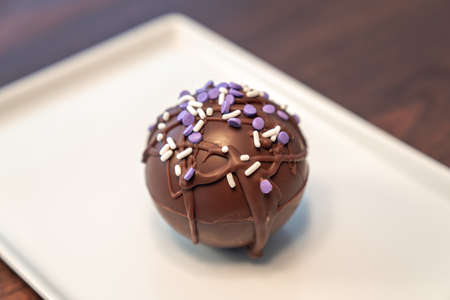 Close up food photograph of a hot cocoa or chocolate bomb covered in drizzled brown chocolate and purple and white sprinkles set on a rectangular plate making a great gift during the winter holidays.の写真素材