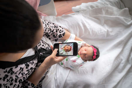 A mother in a pink animal print bathrobe sits on a hospital bed holding her newborn baby girl swaddled in a blanket while taking a photo with her mobile cell phone to share with family and friends.の写真素材