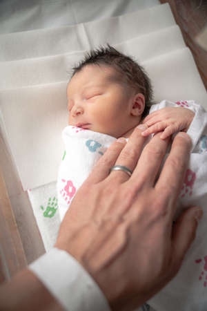 A newborn infant baby girl in a blanket swaddle rests her tiny hand and fingers on her fathers wrinkled hand as she sleeps peacefully.の写真素材