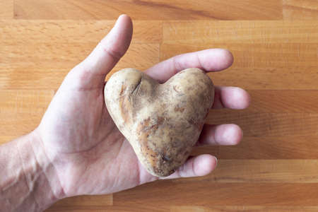 A man holds a heart shaped white potato in his open palm and fingers on a wooden cutting board...