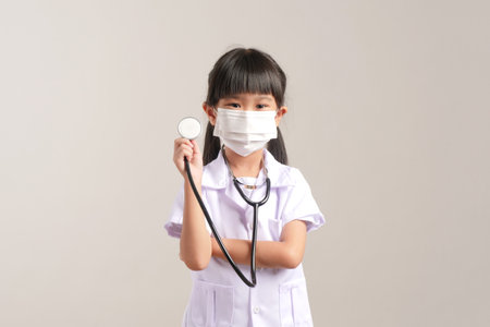 Little asian girl wearing mask and holding stethoscope on gray backgroundの写真素材