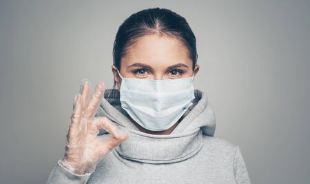 Studio portrait of young woman wearing a face mask and gloves showing okay gesture, looking at camera, on gray background. Coronavirus pandemic, dust allergy, protection against virusの写真素材