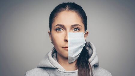 Studio portrait of young woman wearing a face mask, looking at camera, close up on gray background. One half face in mask another withoutの写真素材