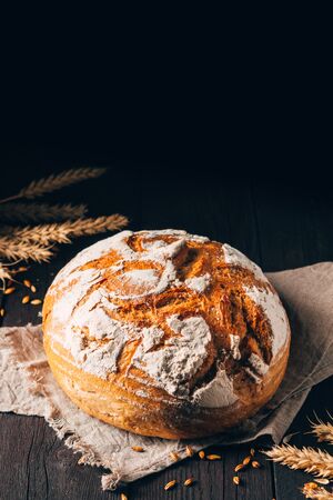Loaf of brown bread with cracks and flour on textile towel on black wooden background