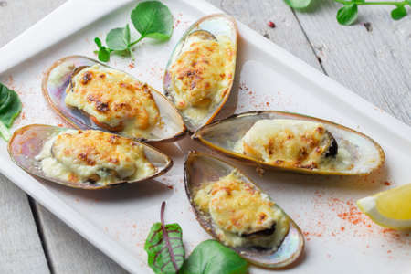 Mussels baked in cheese sauce on a rectangular platter. Decorated with lettuce leaves, spices and lemon wedgesの写真素材
