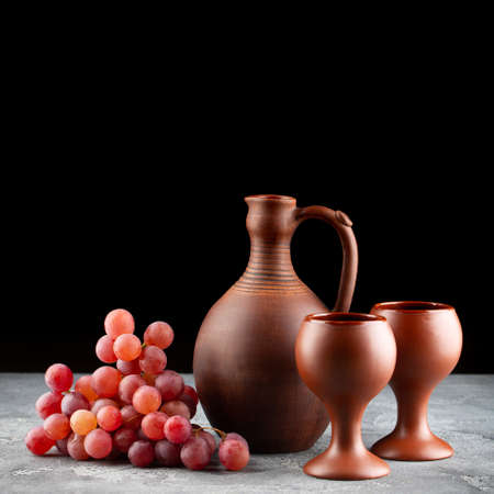 Clay wine jug with two glasses and bunch of red grapes on table on dark background. Copyspace