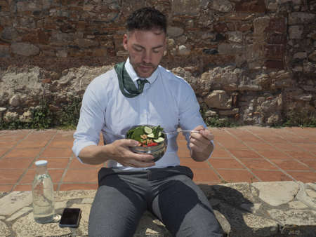 Businessman in tie eats a salad on his outdoor break, he is sitting and holding a bowl with lettuce, tomato, cucumber, Mediterranean food.の写真素材