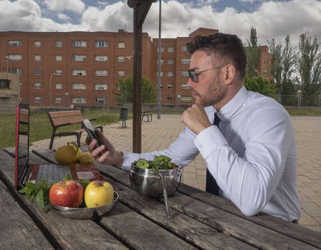 Man eating a salad, with pieces of fruit on the table, he is looking at his mobile phone and working on the computer outdoors.の写真素材