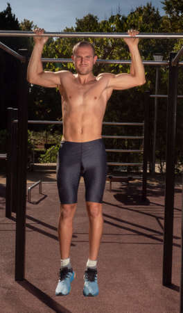 A young man training on a calisthenics bar, wearing sportswear without a shirt. Exercising in a...