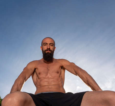 low angle shot. Caucasian man with shaved head and long beard looking at camera, shirtless and with strong abs. background blue sky.の写真素材