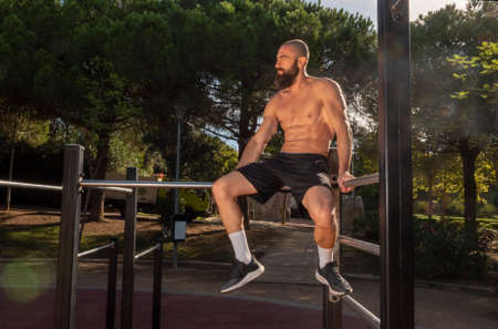 Male sportsman, shirtless, with beard and shaved head, sitting on top of calisthenics bars. Outdoors, in the morning light, in a park with pine trees in the background, looking to his right. horizontal photograph.の写真素材