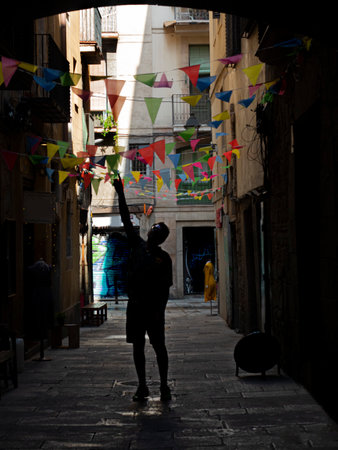 barcelona street with little colored flags decorating the street a person in the backlight is standing trying to touch the flags with one handの写真素材
