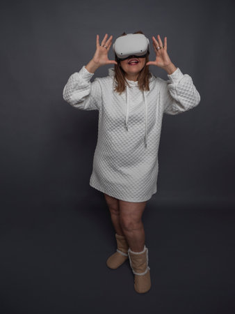 adult woman with virtual reality glasses, she is happy and having fun having fun in virtual reality, she is wearing the virtual reality glasses mask, hands at the height of the mask, full body, comfortable white clothes, gray background.の写真素材