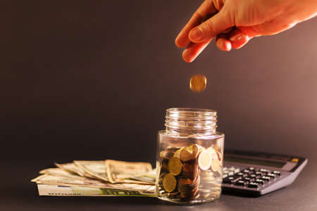 A womans hand lowers a coin into a glass jar on a black background with a calculator. Finance and...