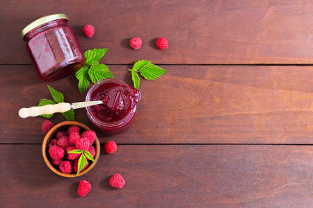 fresh raspberry jam in a glass jar on a wooden table, next to fresh raspberries. concept of homemade...