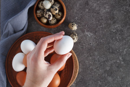 A womans hand holds a chicken egg over a plate of eggs on the kitchen table.の写真素材