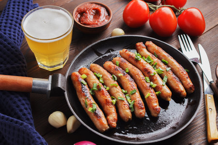 Delicious sausages cooked in a frying pan with vegetables, various sauces and a glass of beer on a wooden table.の写真素材