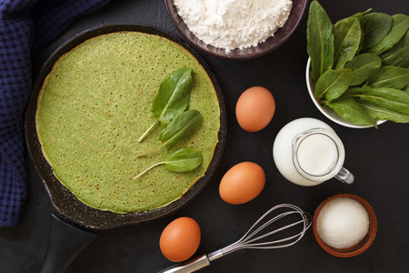 Spinach pancake in a pan on a black background with ingredients: eggs, spinach, milk, flour. Cooking.の写真素材