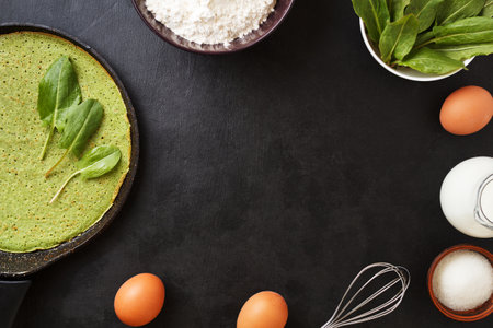 Spinach pancake in a pan on a black background with ingredients: eggs, spinach, milk, flour....