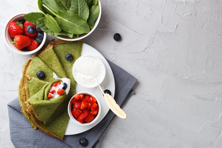 Green pancakes with spinach on a plate with fresh berries and sour cream, on a gray background with textiles. copy spaceの写真素材