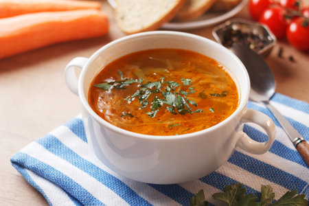 Vegetable soup with potatoes, cabbage, carrots and onions in a white bowl on the kitchen table. Vegetarian food.の写真素材