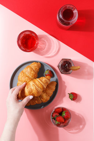 A woman's hand holds a kraussant over a plate with fresh puff croissants, berry juice, jam and fresh berries on a pink and red background.の写真素材