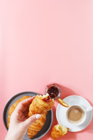 A womans hand holds a croissant over a plate with fresh puff croissants, coffee and berry jam on a pink background. copy space.の写真素材