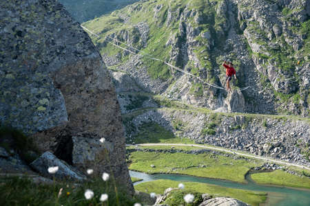 Lago Nero, Pinzolo, Italy - 2020 july 18th: young boy with red hoodie and hat practicing slackline on a rope hanging between two peaks, great height above him, concept of adrenaline and outdoor activitiesのeditorial素材