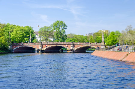Berlin, Germany - April 22, 2018: Banks of the river Spree with citizens enjoying the warm spring day and the Luther Bridge (German: Lutherbruecke) with cast-iron candelabras and obelisks decorated with golden stars in the center of the city.のeditorial素材
