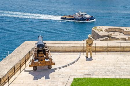 Valletta, Malta - June 4, 2017: View of  Saluting Battery from the Upper Barrakka Gardens, with the guard in uniform and a cannon and a passing ship in the Grand Harbourのeditorial素材