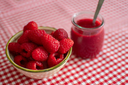 Small bowl with fresh raspberries next to a glass with raspberry jam. Cute crockery.