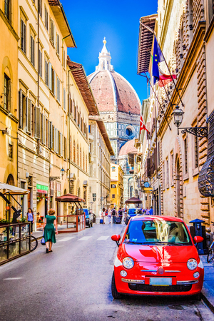 Red small car is parked in narrow street of Florence with view of Basilica Santa Maria del Fiore, famous retro vintage automobile is symbol of Italy, Florence, Tuscany, Italy.の写真素材