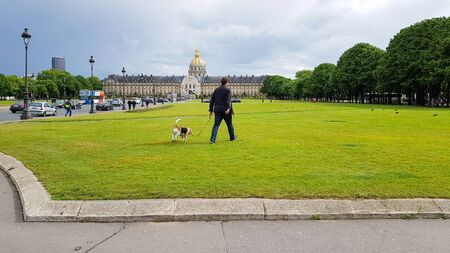 PARIS, FRANCE - MAY 9, 2019: Parisian man walks with dog on green lawn fields near Disabled House, Les Invalides Museum with golden dome, Paris, Franceのeditorial素材