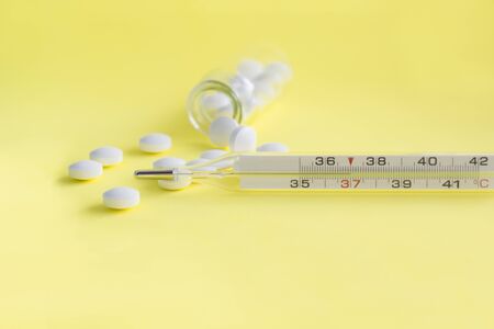 Glass mercury thermometer and white medical pills, tablets from glass bottle on yellow background. Pharmacy theme, treatment and prevention of virus. Concept of health in season of colds.の写真素材