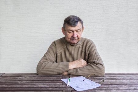 Portrait of elderly man sitting at table with white sheet of paper, pen and glasses.の写真素材
