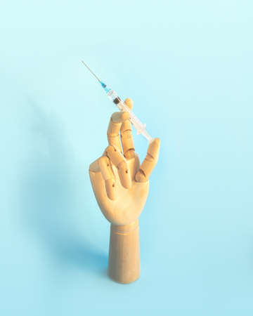 Wooden human hand with syringe with vaccine for disease on light blue background. Doctor holds syringe with medical injection. Health care and disease prevention concept. Vertical photo.の写真素材