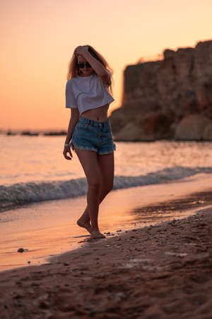 Girl walks along beach of sea at beautiful sunset above horizont toning in calming coral color. Trendy creative design in color of 2022. Female feet walking along coastline on calm summer eveningの写真素材