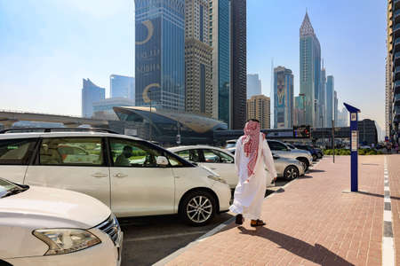 Dubai, UAE - November 27, 2021: View of iconic red telephone booth and Emirates towers buildings similar to London style in Dubai. Man in national dress, United Arab Emiratesのeditorial素材