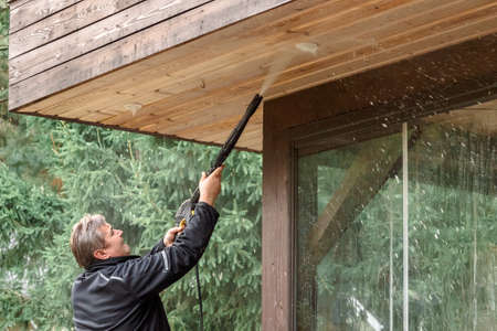 Man cleaning walls and floor with high pressure power washer. Washing terrace wood planks and cladding walls. High quality photoの写真素材