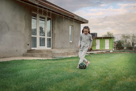 Cute little girl playing football with soccer ball on green lawn in backyard of house. Child kicking...