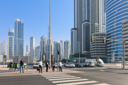 UAE, Dubai - November 27, 2021: People walk along pedestrian crossing in Business Center in Dubai. Men in suits and medical masks rush to work against backdrop of huge skyscrapers. High quality photoのeditorial素材