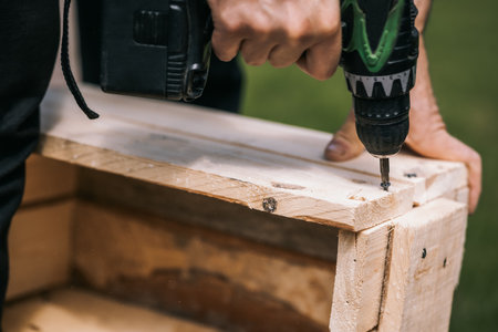 Man is screwing screw into wooden box with cordless screwdriver. Product repair with professional tool electric screwdriver. Close up. High quality photoの写真素材