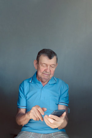 Elderly man dials number in smartphone from home. Old senior sends sms on mobile phone. Vertical photo. High quality photoの写真素材