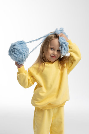 Pretty little girl in yellow clothes plays with ball of blue thread. Child model with long blond hair sits in studio on light background.の写真素材