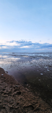 Spring landscape. Panoramic view of beautiful dawn on bay. Cumulus clouds over water in bright light. Ice, snow and rocks on coastline. Rising rays of sun are reflected in sea. Vertical photo.の写真素材