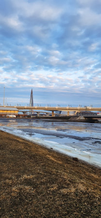 Serene sunrise over a partly frozen river with modern buildings in the background. The sky is filled with clouds, a pathway runs alongside, offering a peaceful morning scene.の写真素材