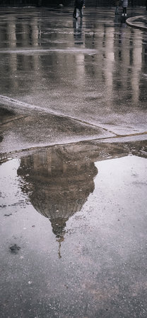 Rainy day city scene with architecture reflected in a puddle. People walk past, suggesting a quiet urban atmosphere. The photo captures a blend of weather and history.の写真素材