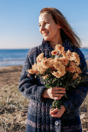 Female portrait. Beautiful young girl with long dark hair walking with bouquet of flowers along beach on sunny autumn day. Vertical photo. High quality photoの写真素材
