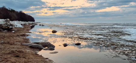 Sunrise over a tranquil coastline with icy patches and smooth stones along the sandy shore. The sky reflects in the water, creating a serene and peaceful atmosphere.の写真素材