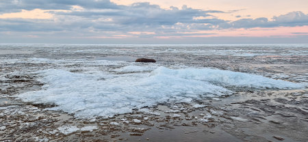 Sunrise over a tranquil coastline with icy patches and smooth stones along the sandy shore. The sky reflects in the water, creating a serene and peaceful atmosphere.の写真素材